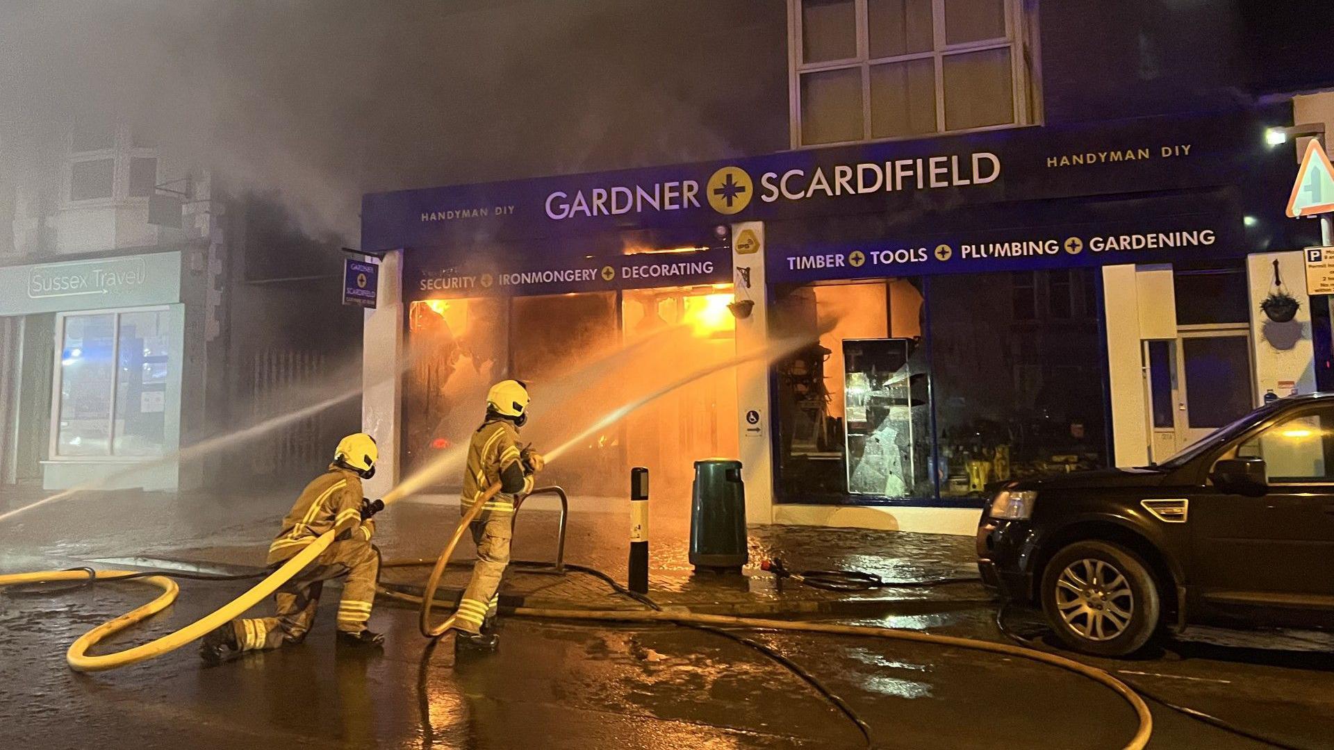 Two firefighters spraying water at a building. The building has a blue shop front and flames on the inside. 