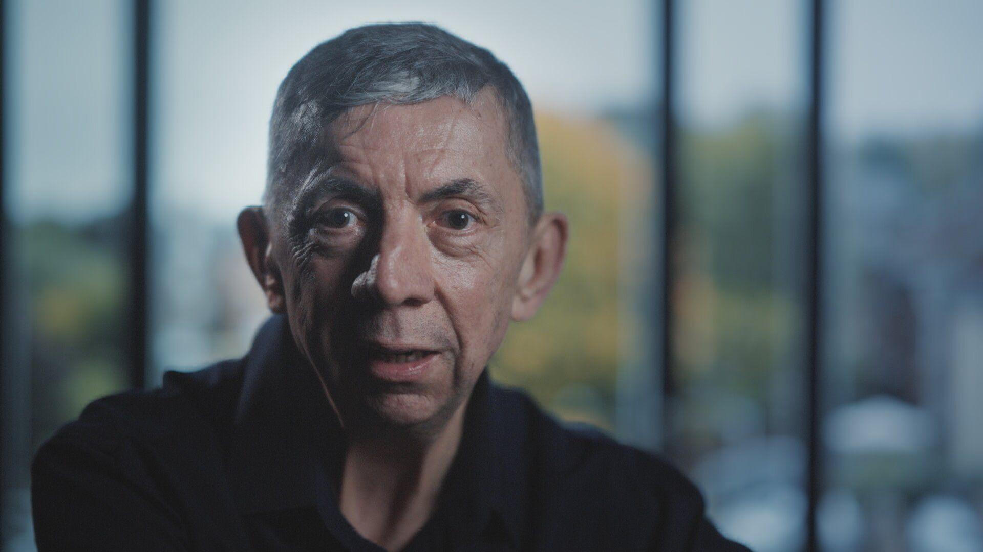 Gary Hales is looking at the camera. He has a slim face and is clean-shaven. He has dark eyes and short, grey hair. He is wearing a dark-coloured top