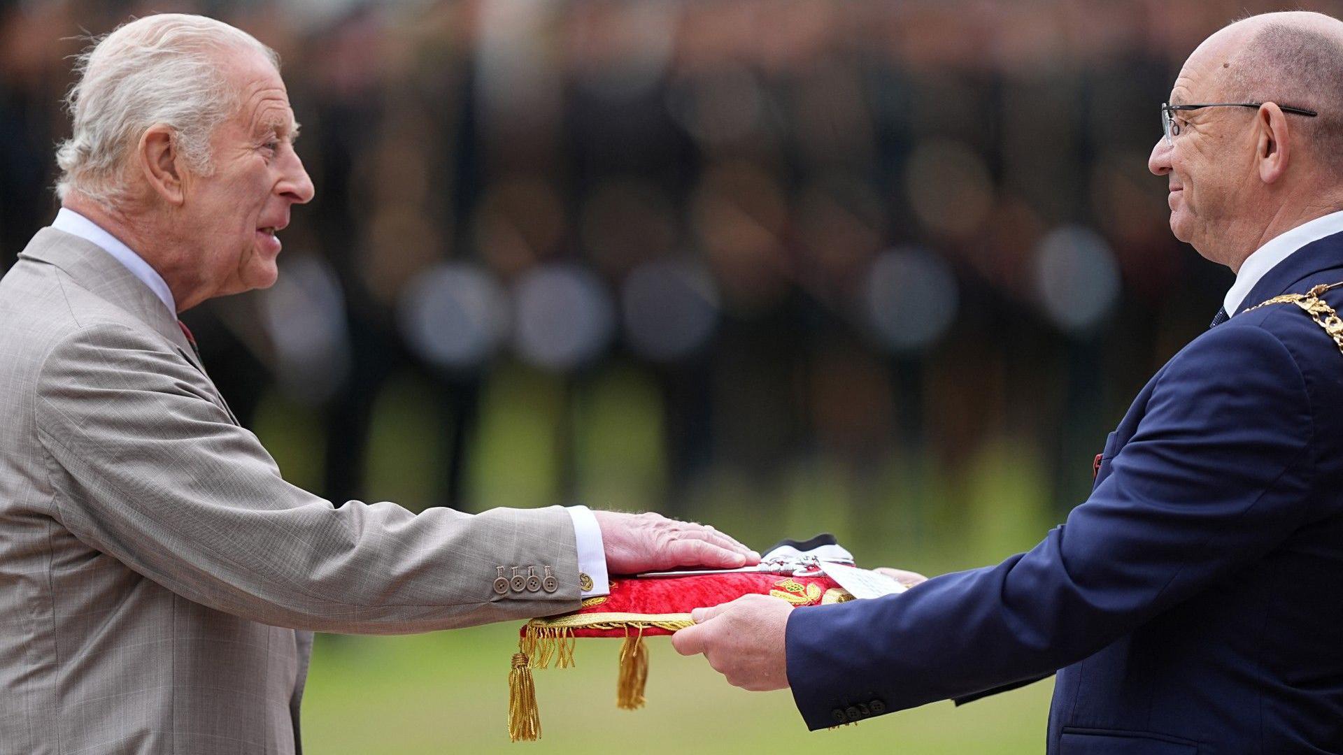 King Charles III wearing a grey suit and white shirt places his hand on a red cushion held by the Lord Provost of Edinburgh Robert Aldridge, wo wears glasses and a navy blue suit. In the background, lines of soldiers in military uniforms are blurred.