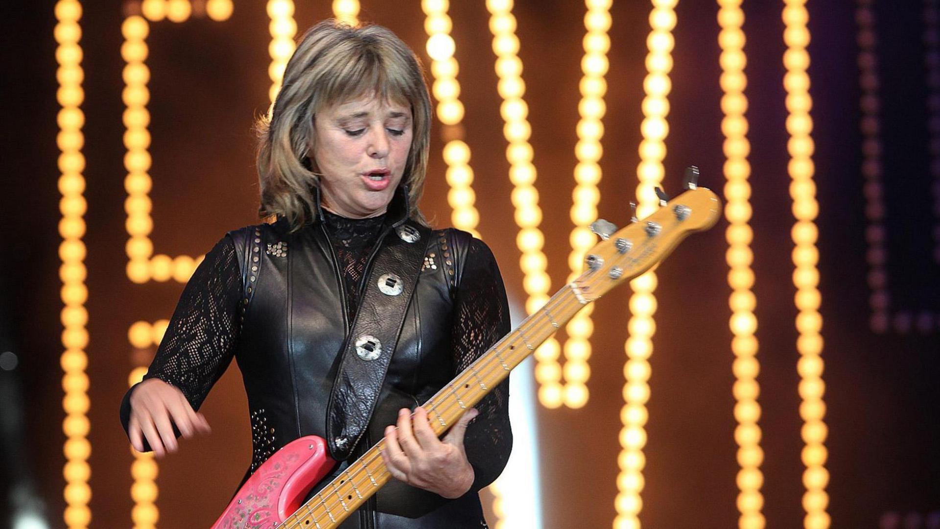 Suzi Quatro wearing a black leather outfit while playing a pink guitar on stage.