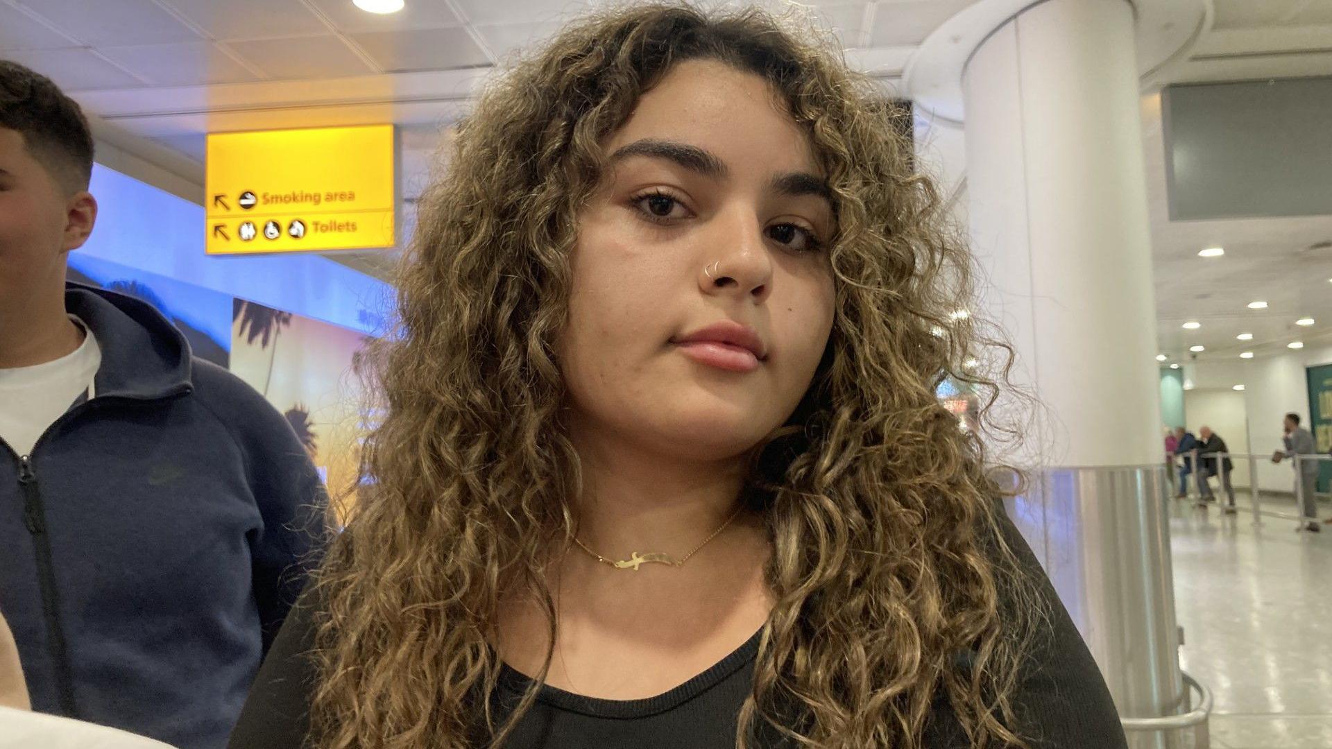 A photo of Lara, a teenager with curly hair and a dark top, at Heathrow Airport. She is looking into the camera.