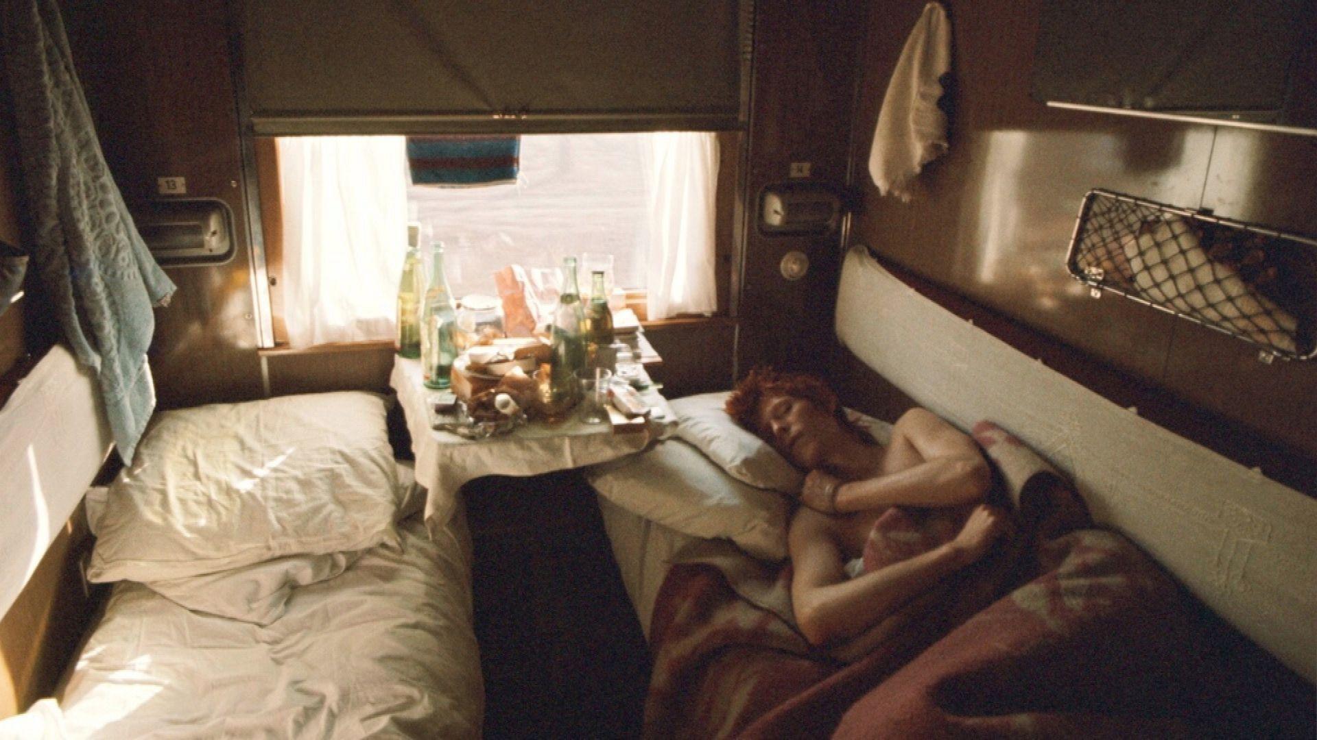 Geoff MacCormack photographs David Bowie while asleep on the Trans-Siberian Express. There star is in a carriage with two single beds. He is sleeping next to a table filled with bottles of drinks.