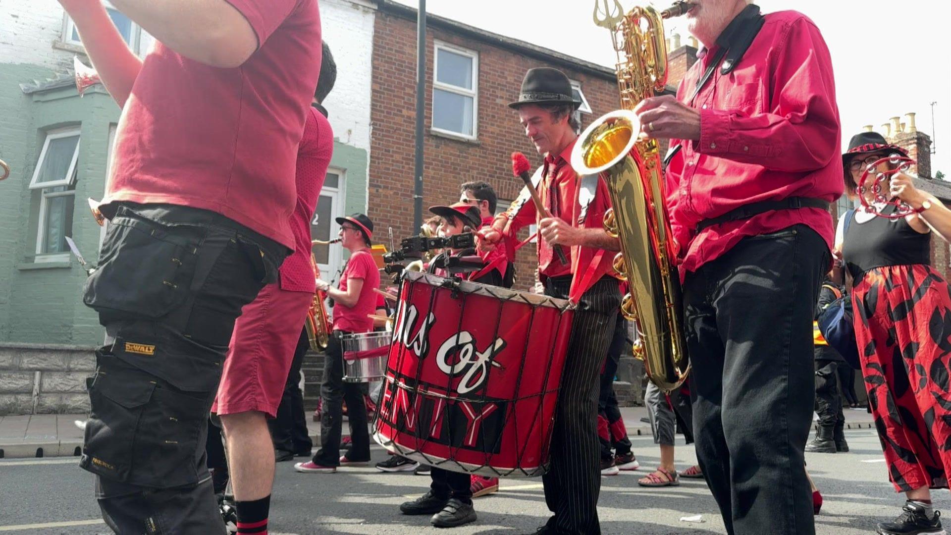 A band, complete with a drummer and saxophonist, marching. They are wearing black trousers and red shirts