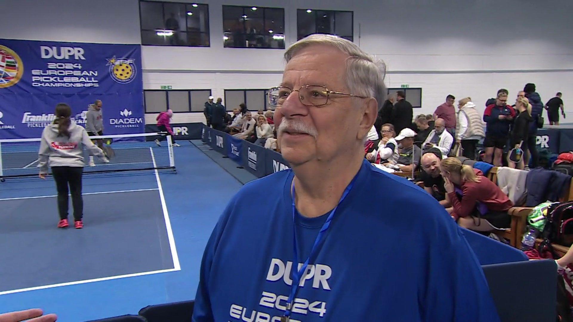 President of the European Pickleball Federation Frank Arico being interviewed. He is looking away from the camera. Players could be seen on the court and in the seats behind him.