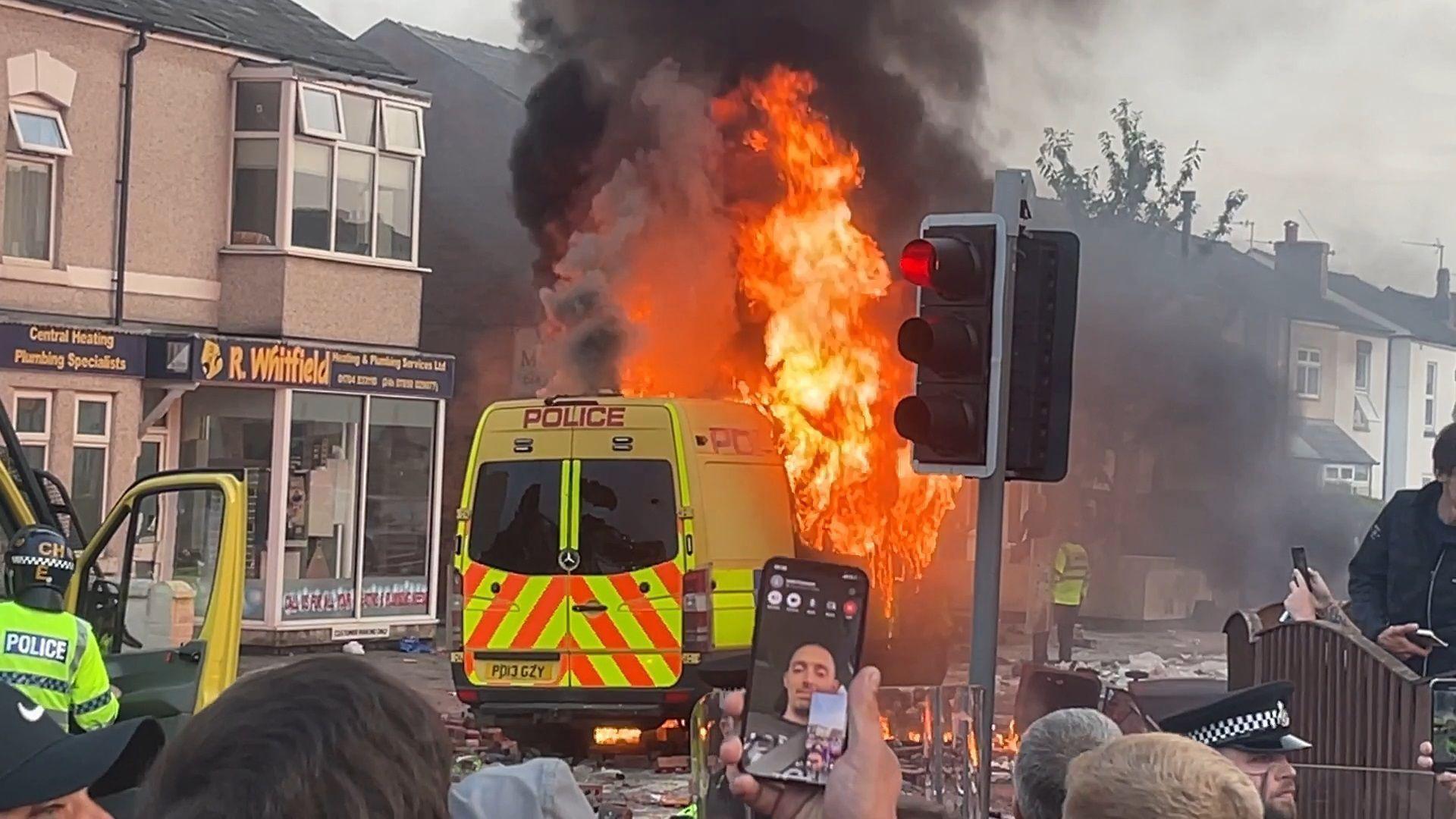 Large orange flames engulf a yellow Merseyside Police van on the debris littered streets of Southport as a crowd watches on, some filming on their mobile phones. 