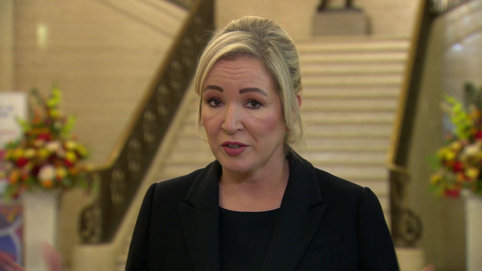 Michelle O'Neill in black jacket and black top. She is standing in front of a staircase. There are flowers behind her. She has blonde hair.