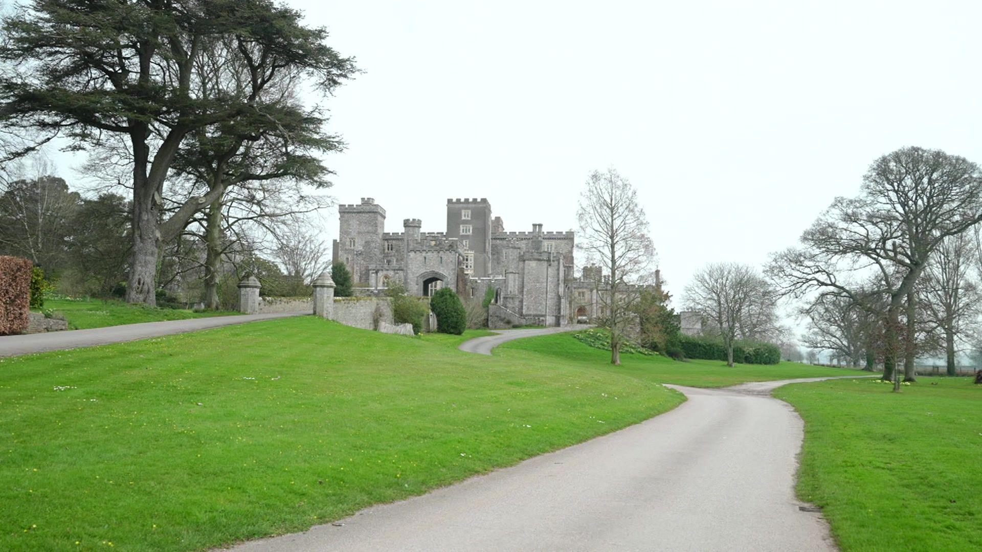 A photo which shows Powderham Castle in the distance. The castle is made of grey bricks and has turrets. In the foreground is green grass as well as concrete road leading up to the castle. There are trees surrounding either side of the castle. 