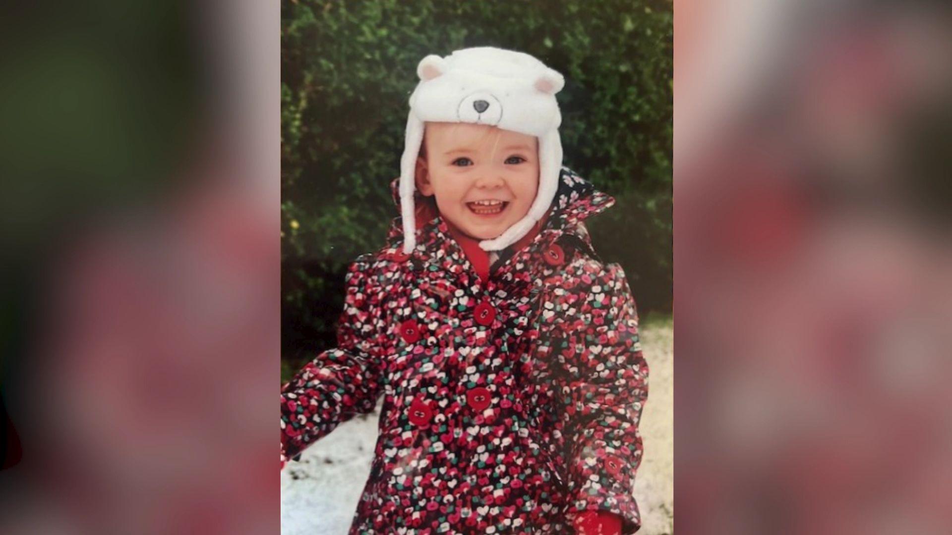 Emily as a toddler, she is outside in the snow wearing a red floral coat and a white polar bear hat. She is smiling and has blonde hair. 