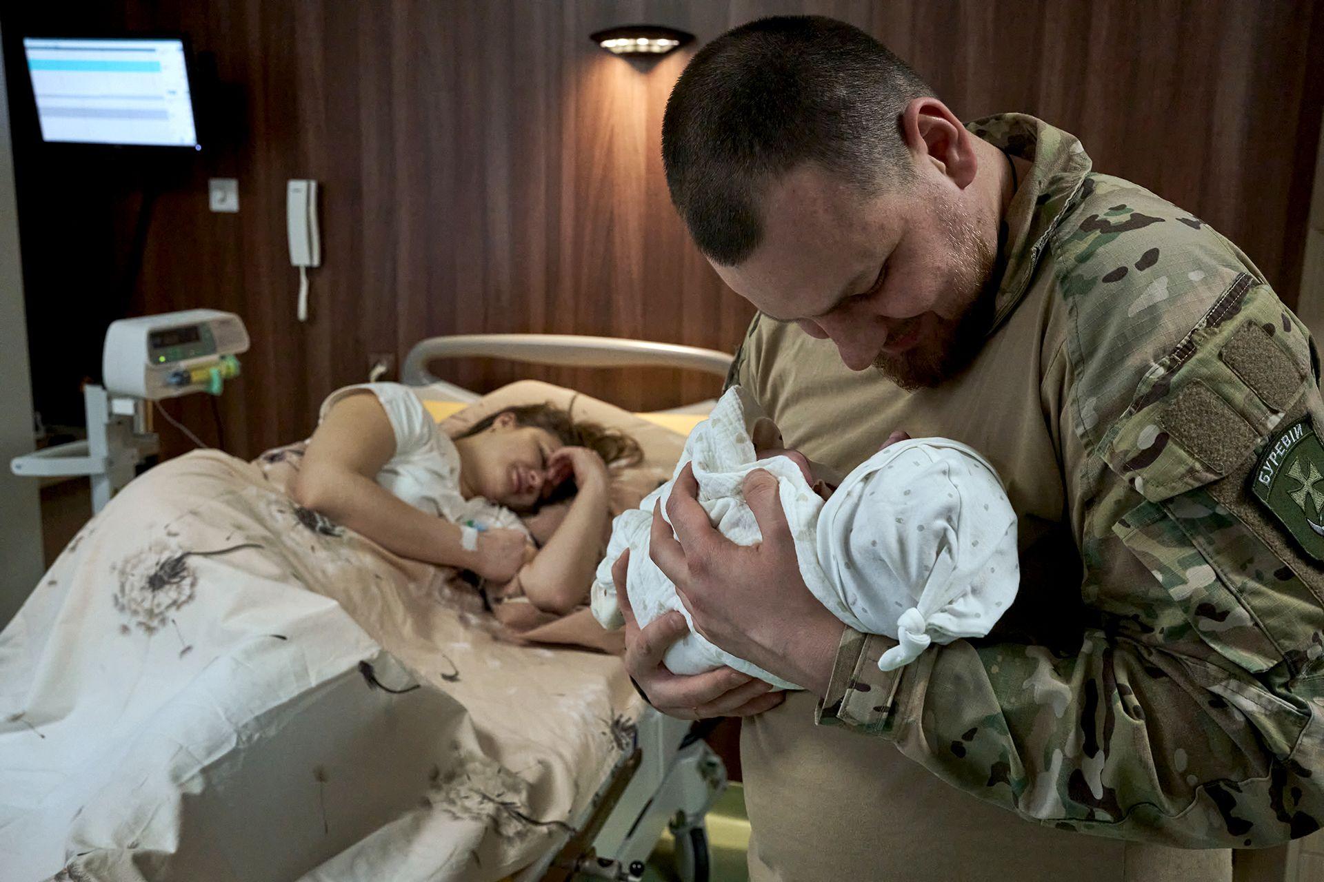 Dmytro looks down at his baby daughter as he holds her in the delivery room with his wife lying on the hospital bed after giving birth; Dmytro wears military uniform and the baby is wrapped in white, taken at the KNP Perinatal Centre in Kyiv, Ukraine on 23 March 2024.