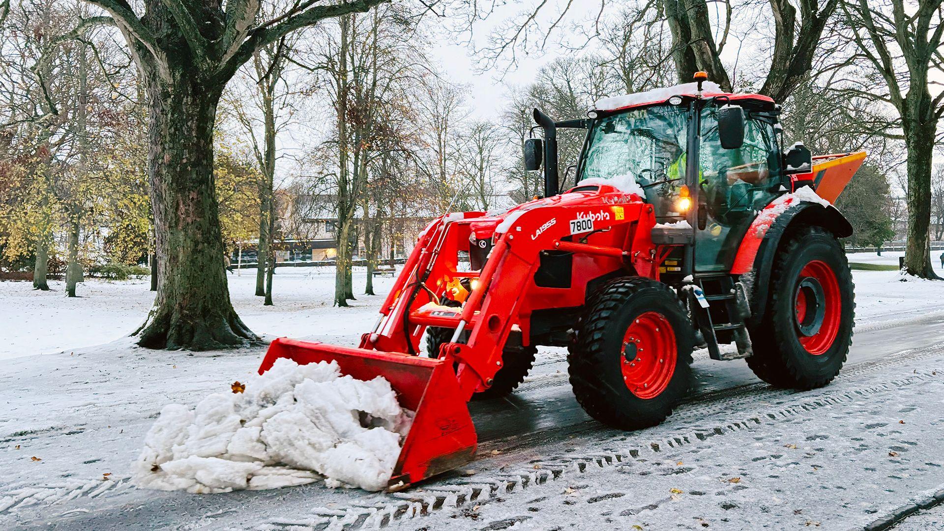 A snow plough clears snow in a park, it is orange and stands out amid the white snow.