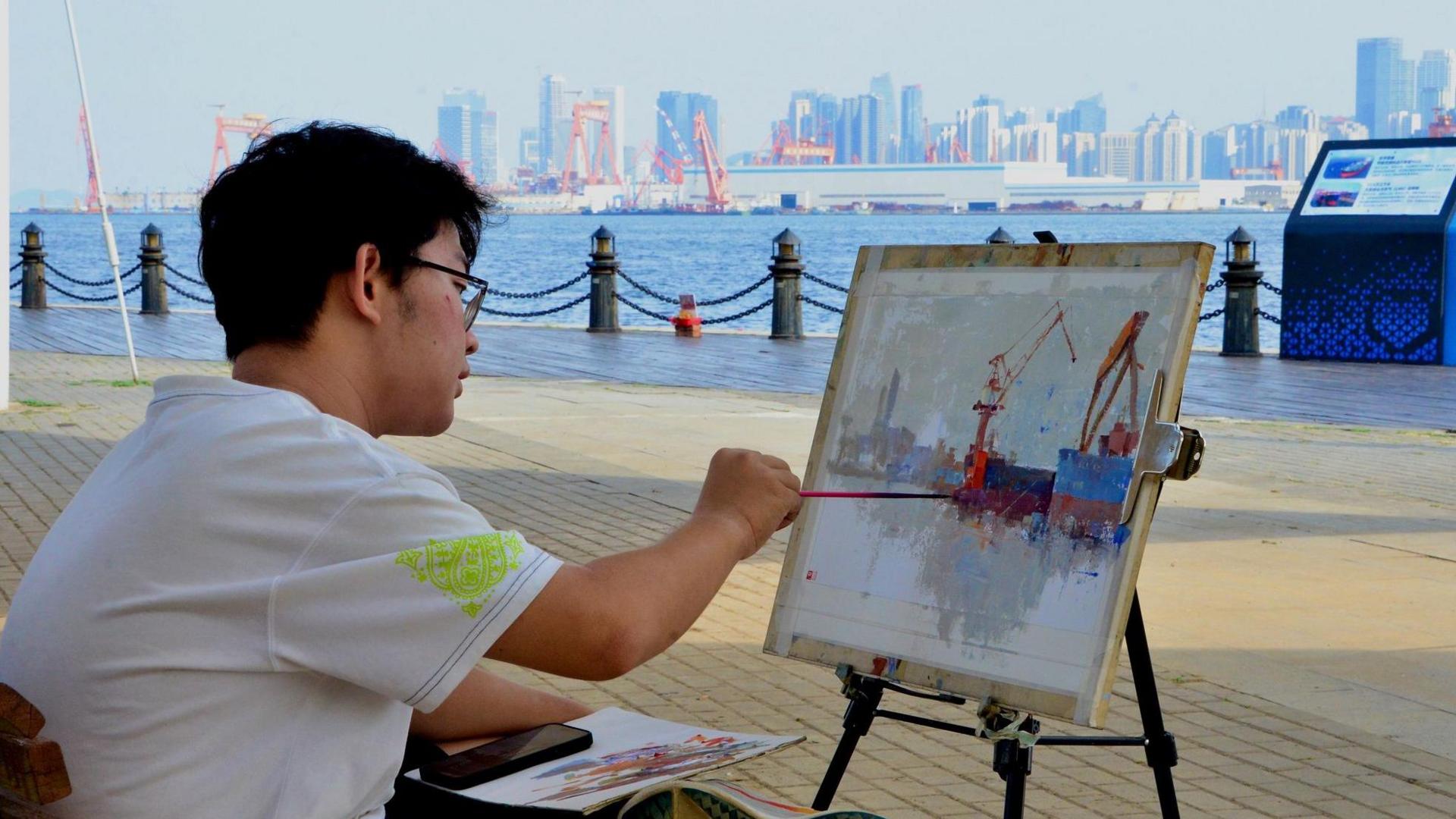 A young man is painting a view of the Dalian shipyard in watercolours. He is sitting across the harbour from it, in a white shirt and black shorts, with an easel in front of him.