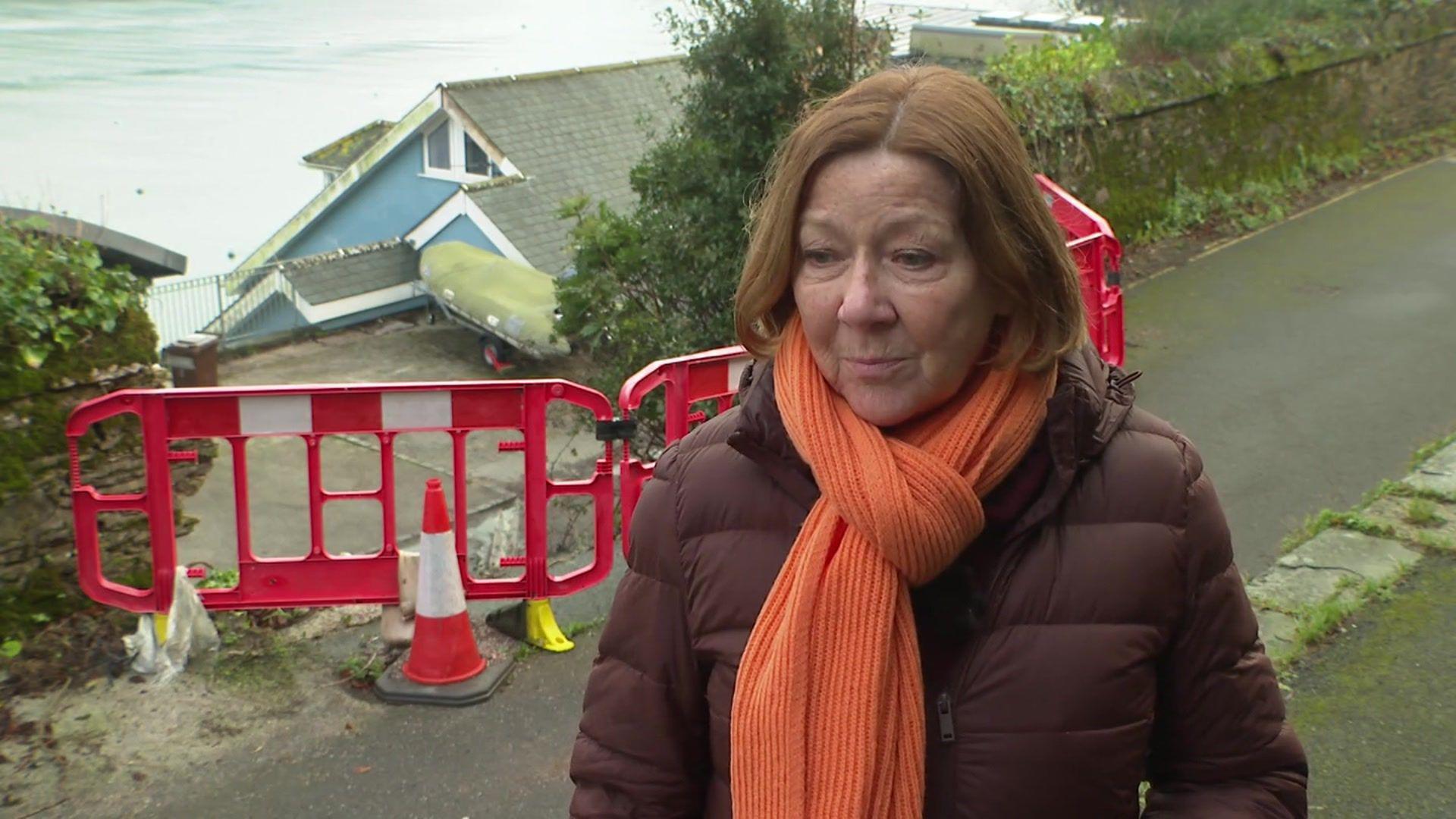 Lindsay Ellwood looking to the left wearing a dark purple coat with an orange scarf. She is stood in front of the barriers surrounding the landslip.