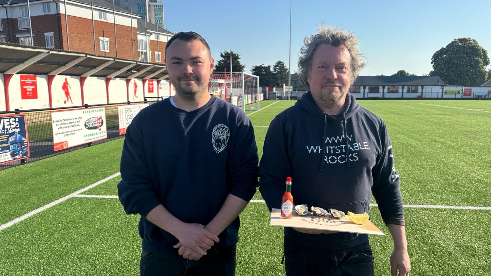 Two man in dark clothing stood on a football pitch. They are facing the camera, the man on the right is holding a wooden board with oysters on