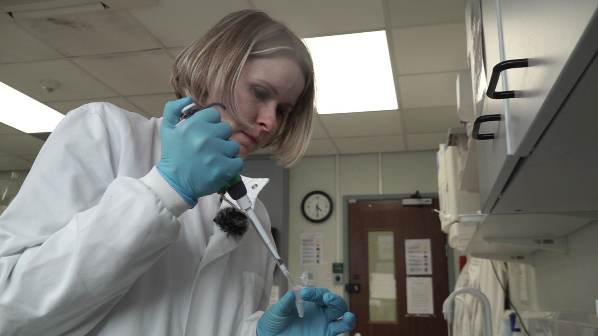 Dr Magna is wearing a white lab coat and blue gloves. She is in a lab and is holding lab equipment.