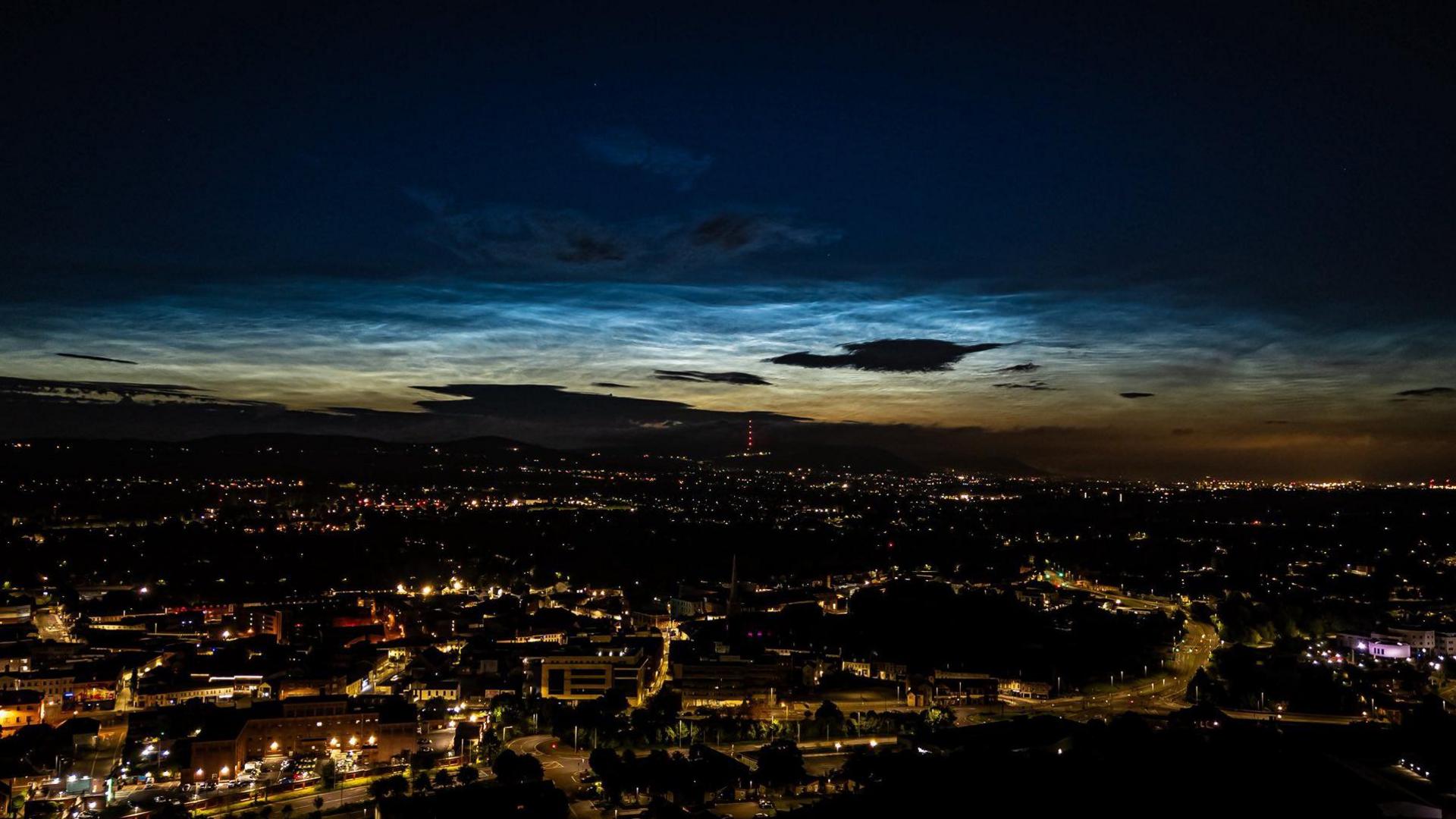 An aerial view of a city is cast mostly in darkness but is faintly illuminated by street lights and a slight strip of sun in the distance. There is a patch of scattered clouds in the sky. 
