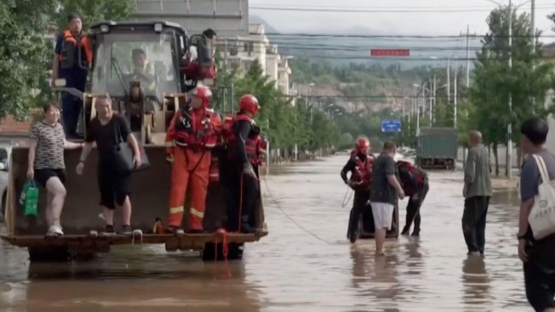 People stand on an excavator vehicle's front loader bucket in the middle of a flooded road.