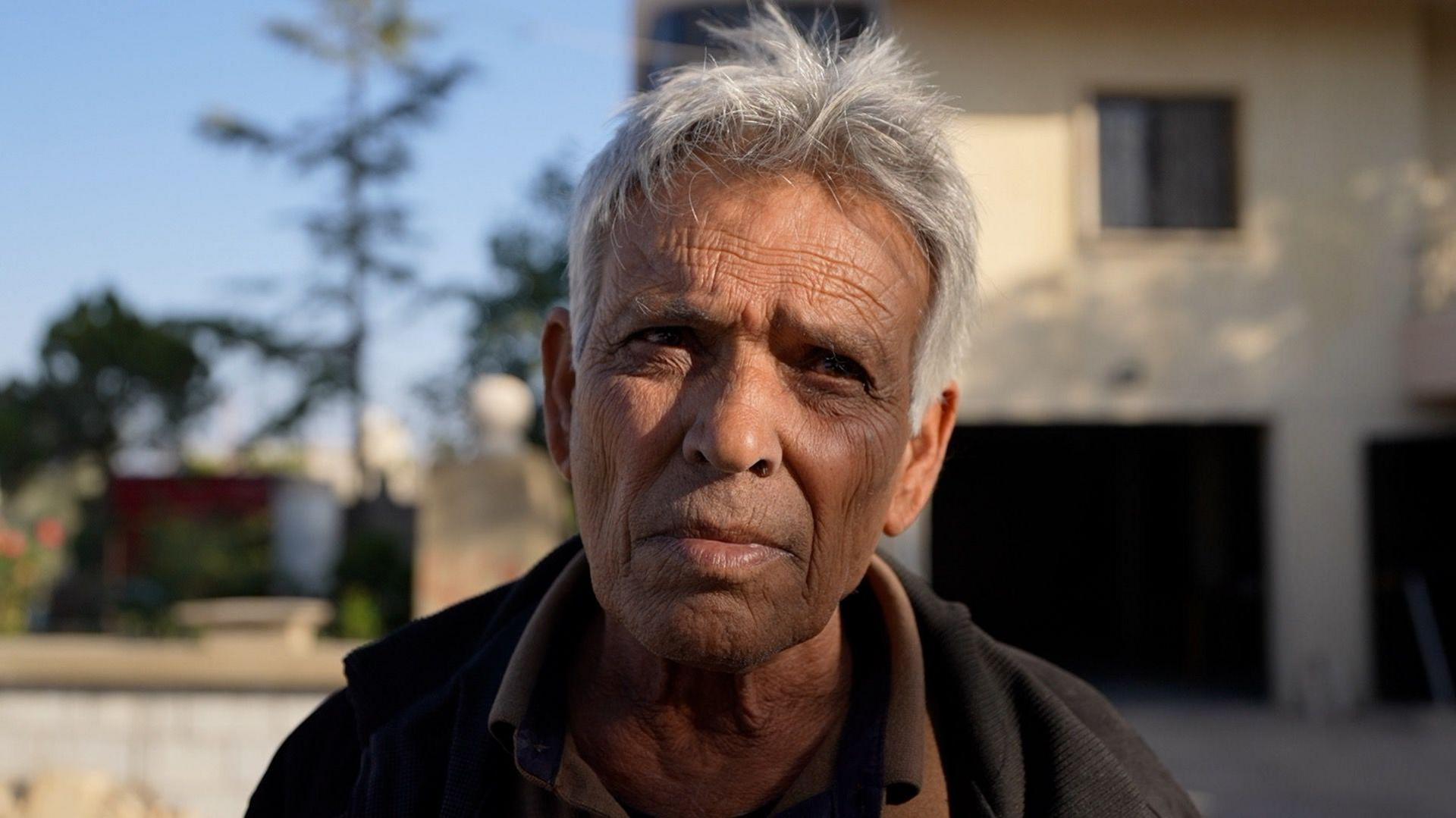 An older man with a weathered face and grey hair looks into the camera.