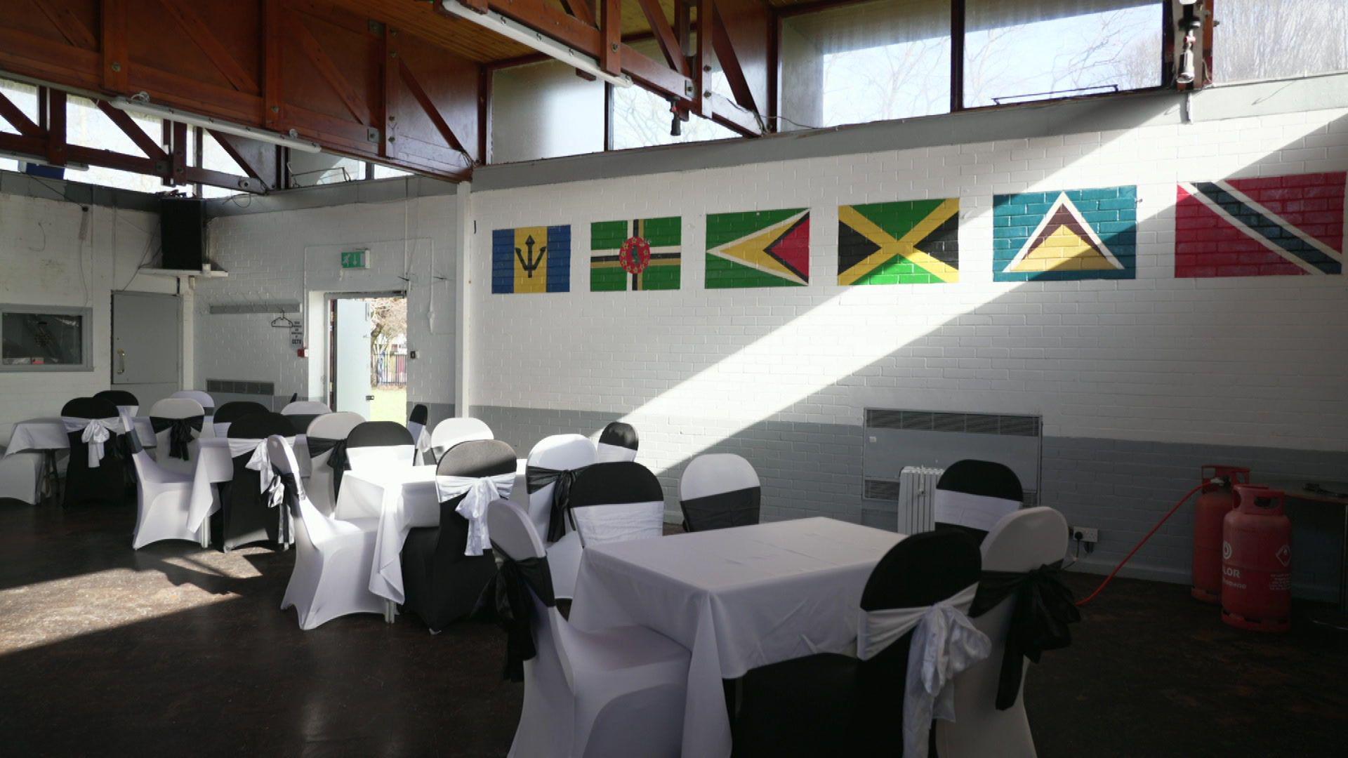 Inside the community hall where tables with white table cloths and chairs with white covers are located. Caribbean flags are painted on the walls including: Barbados, Dominica, Guyana, Jamaica, St Lucia and Trinidad and Tobago