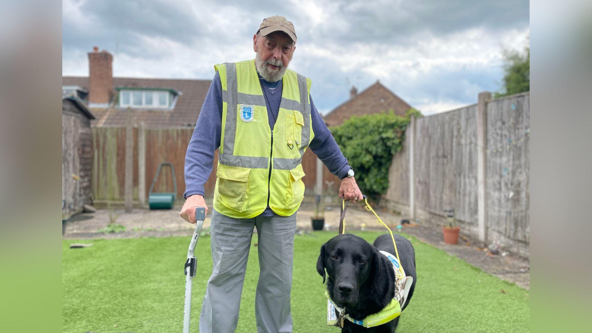 A man with a walking stick in one hand, and a lead for his black labrador guide dog, Hero, in the other