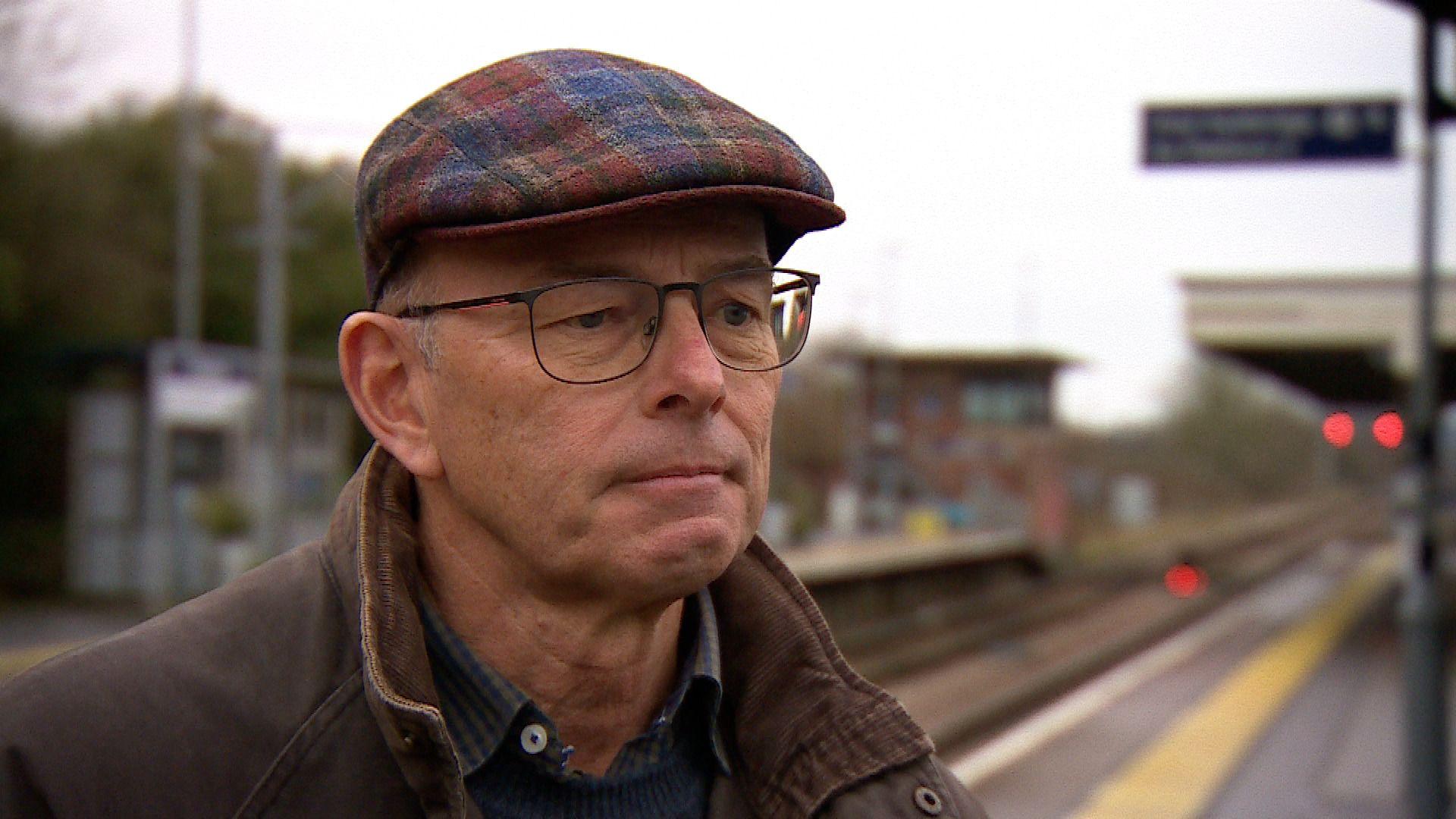 Nick Hurrell is wearing a tartan flat-cap and glasses. He also has a blue jumper and brown coat on. Behind him out of focus is a train platform and railway line.