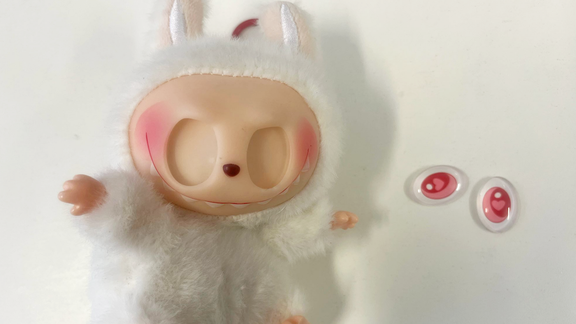 A white fluffy Labubu doll with empty eye sockets. It's large pink eyes sit next to it on the table.