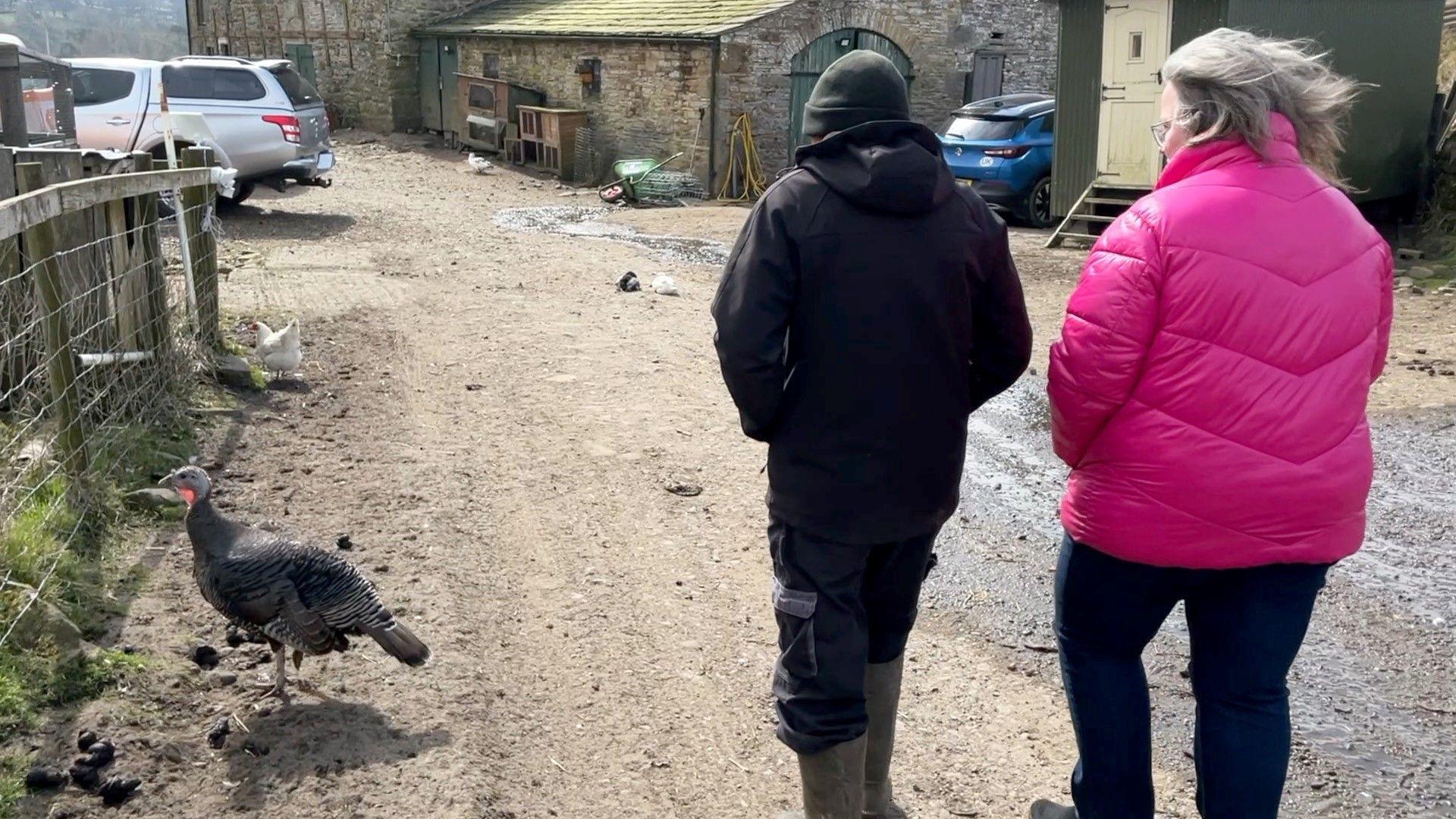 Tony Johnson and his wife Ute walk along a stone track wearing coats, there is a turkey to their left.