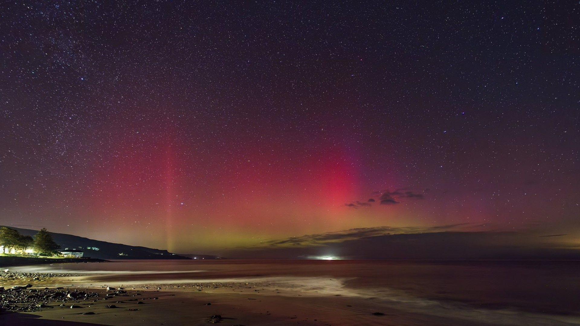 A landscape photo taken at night time from a sandy beach. The sky is full of small, bright stars with flashes of red and pale green lights.