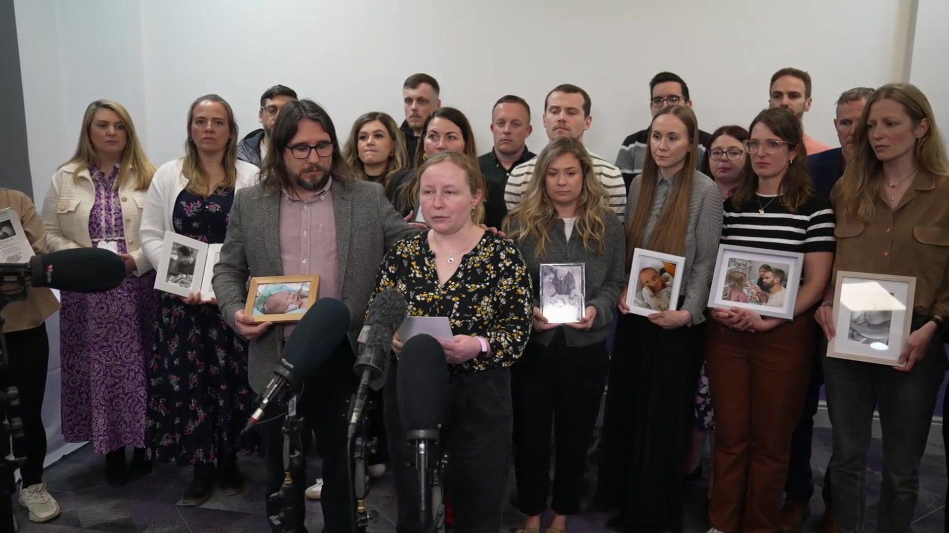 A group of people stand closely together indoors behind several microphones, some holding framed photographs and listening as one person reads from a sheet of paper.