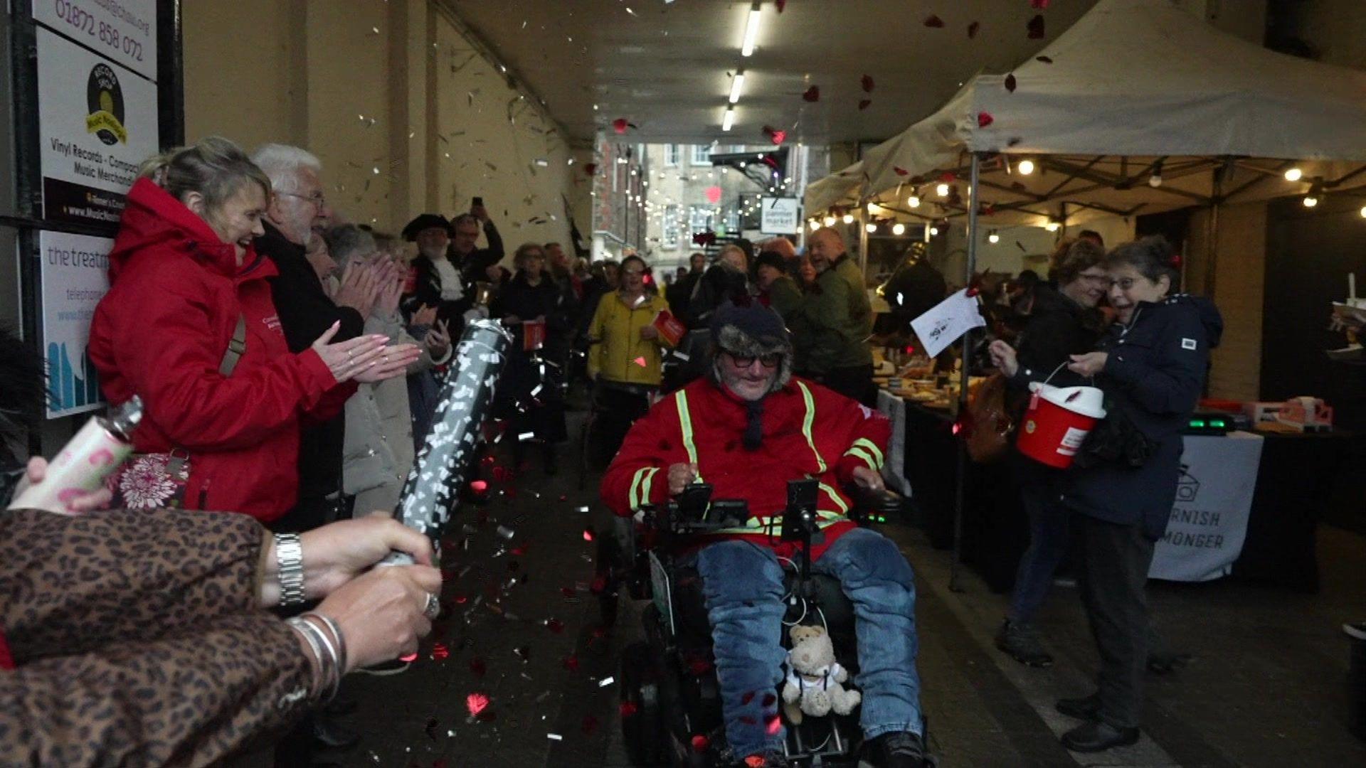 Steven Webb crosses the finish line in a wheelchair as supporters surround him, cheering and setting off streamers