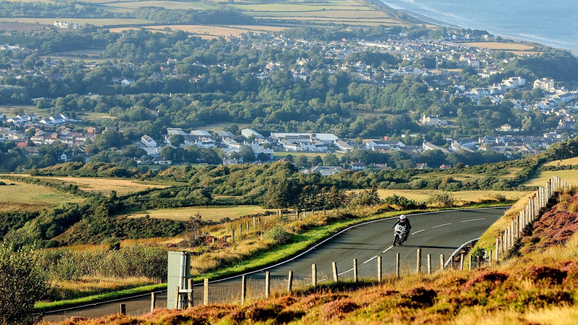 A motorcycle on a grey road on a hillside which has yellow grass in fields along side green trees. In the distance you can see a town made up of houses and other large building near the sea.