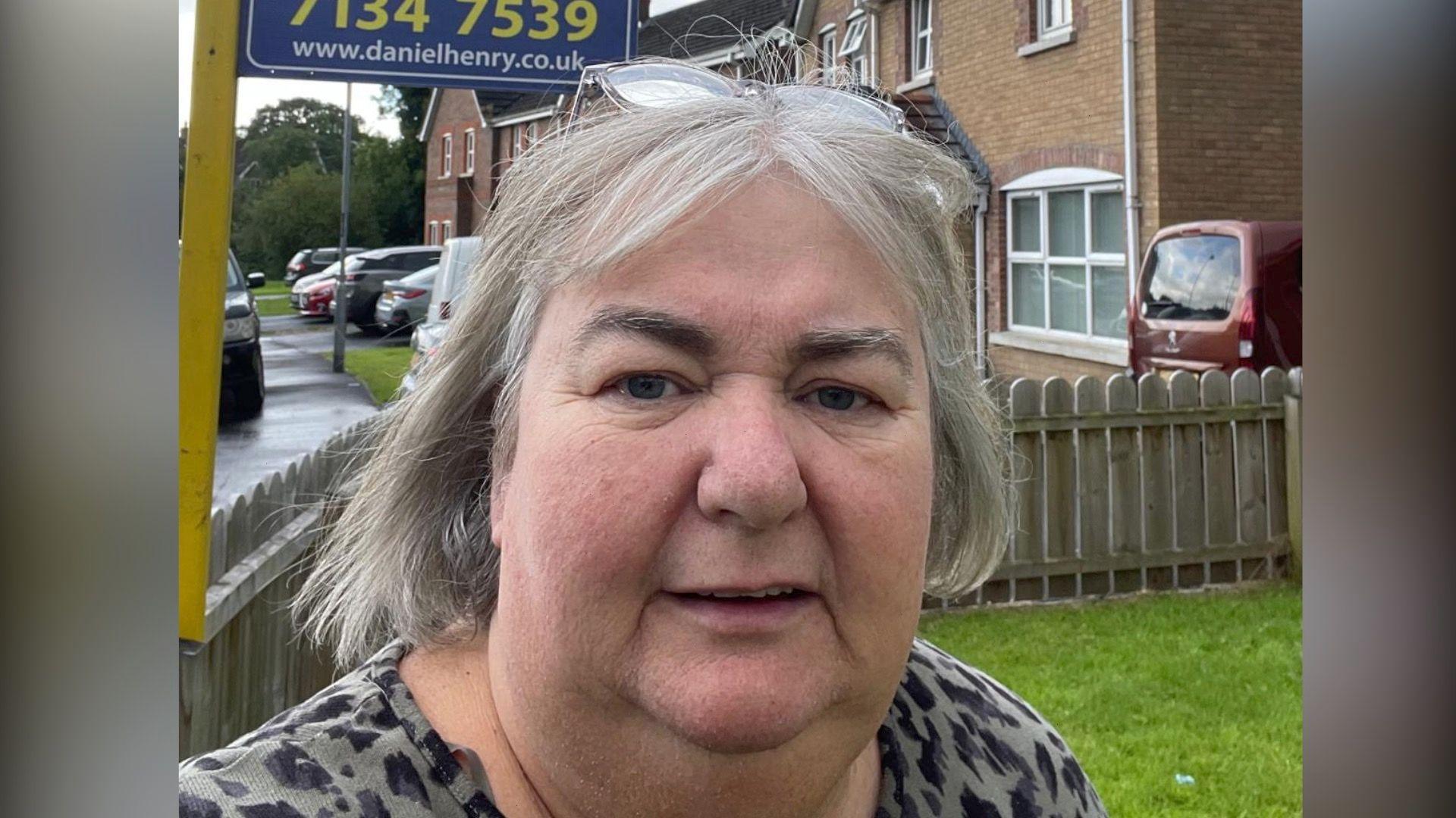 Catherine Cooke takes a selfie standing in her garden, a for sale sign can be seen in the background, as can a number of her neighbours homes