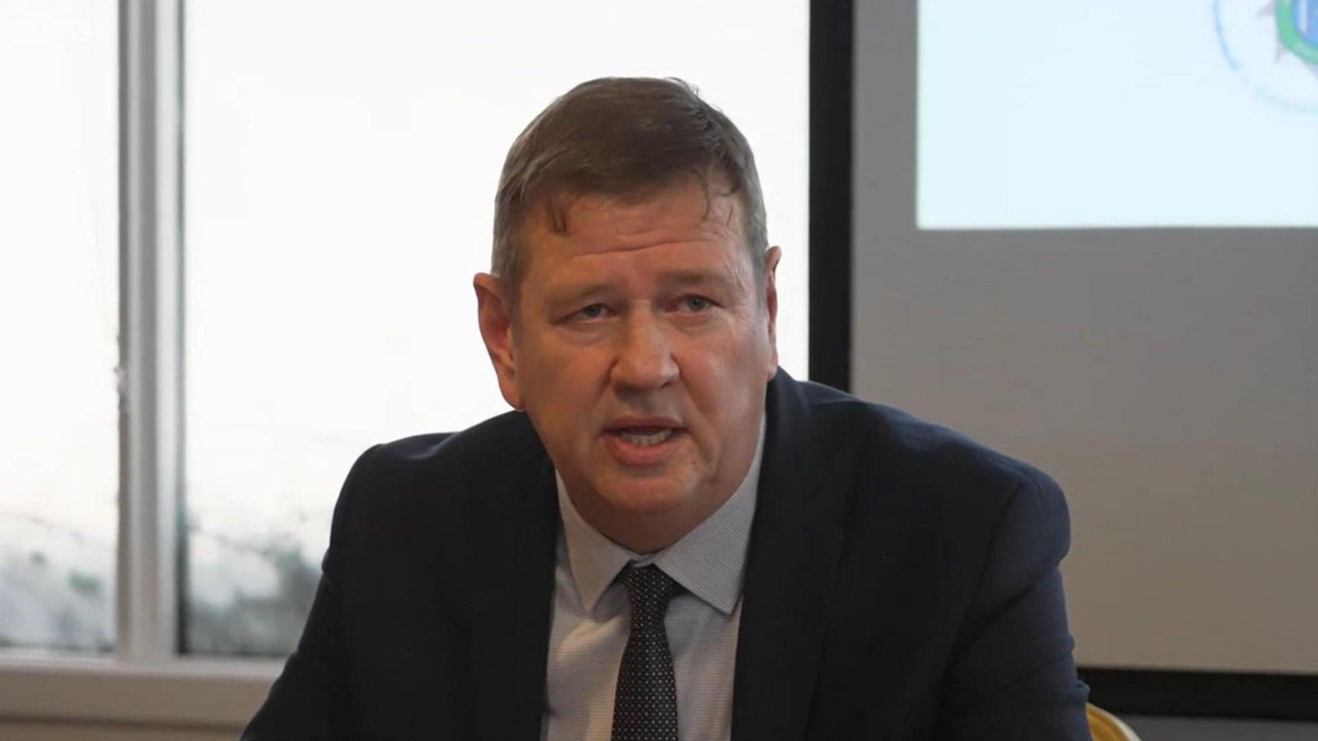 Mark Baker, wearing a dark suit jacket over a grey shirt and tie, speaks to the media at a press conference 