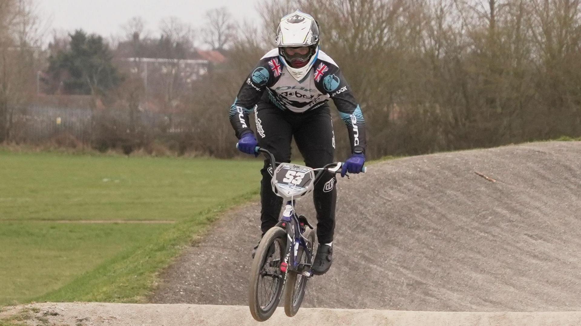 Jared Hill rides his BMX bike over a jump on his home circuit in Norwich.  He is wearing a white, black and sky blue racing suit and white helmet.