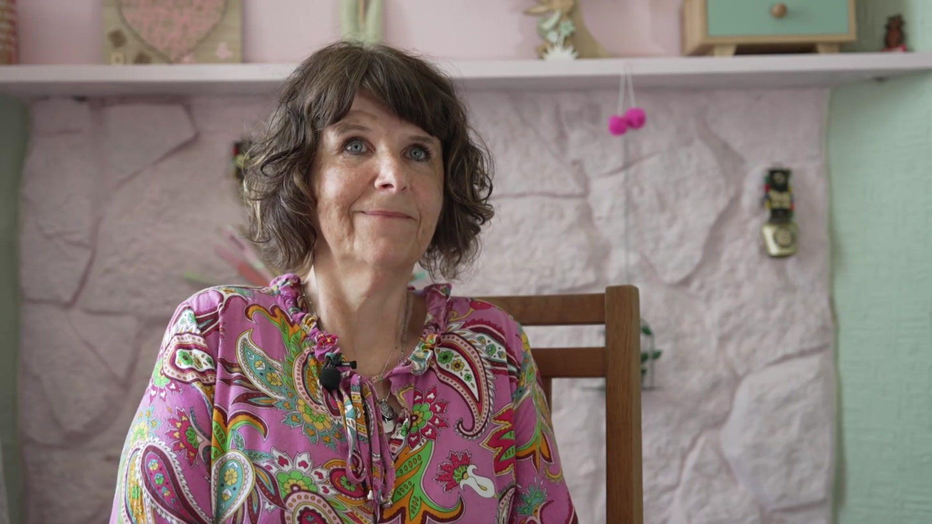 Tiffany is sitting on a wooden chair, smiling at the camera. She is in front of a pale pink and green wall. Her brunette hair is in a short bob and she is wearing a pink top with colourful swirls and shapes on.