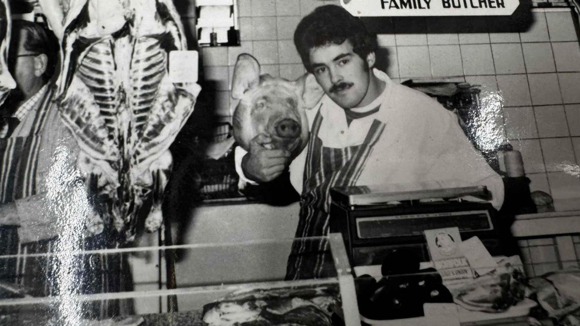 Geoff Hughes with thick hair and a moustache holds a pig's head at a former workplace in a black and white photo.