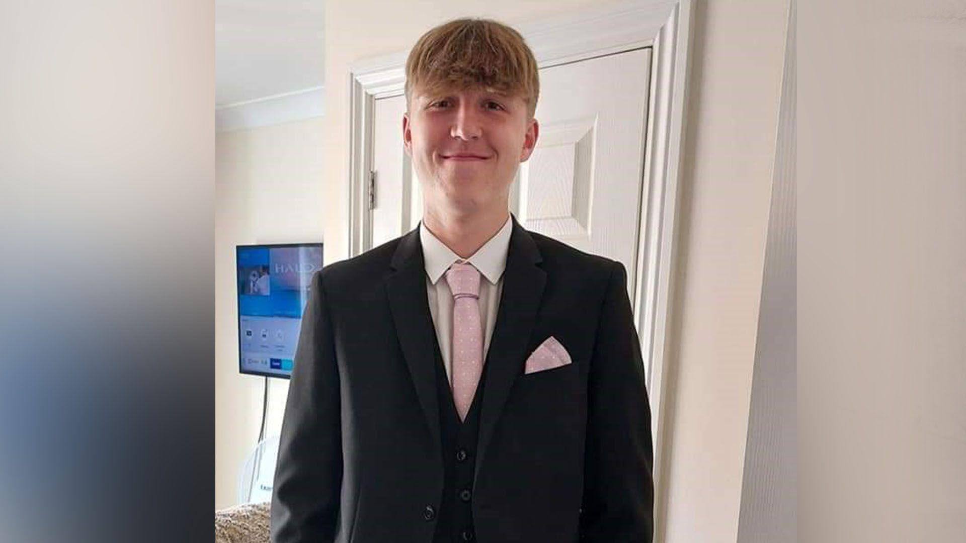 18-year-old Alfie Gray wears a suit with a pink tie and is smiling down the camera.