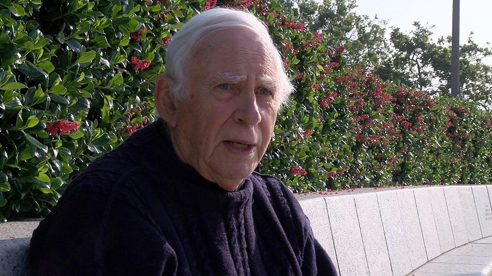 David sits on a white granite curved bench seat. He has short white hair and is wearing a navy blue knitted jumper.