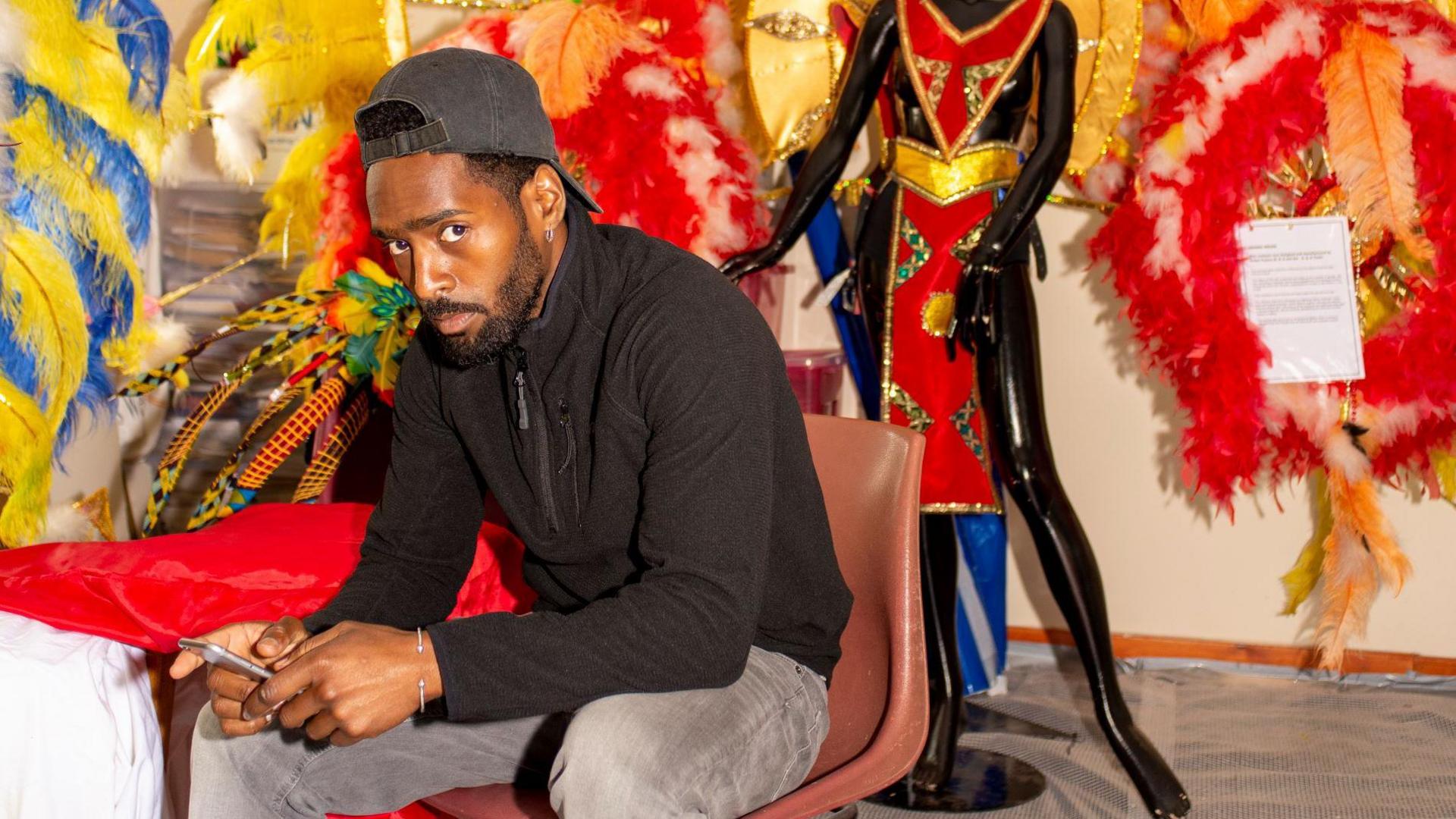 Mr Charles-Kelly sits on a brown chair and holds a phone between his hands as he looks at the camera. He is wearing grey jeans and a black fleece. Red, yellow and blue feathers adorn the wall behind him and a mannequin in a red and gold garment is positioned behind him. 