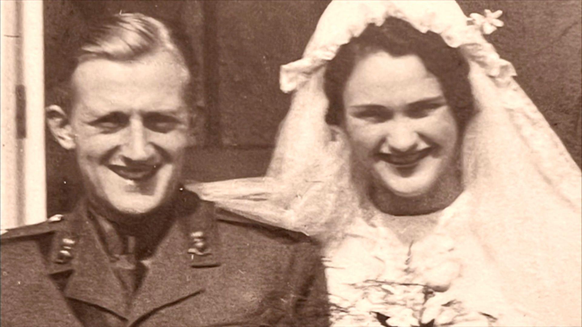 A sepia-colour archive photo of Margaret and Rex stood together on some steps outside a building on their wedding day. She is wearing her wedding dress and holding flowers, while he is smiling in military uniform.