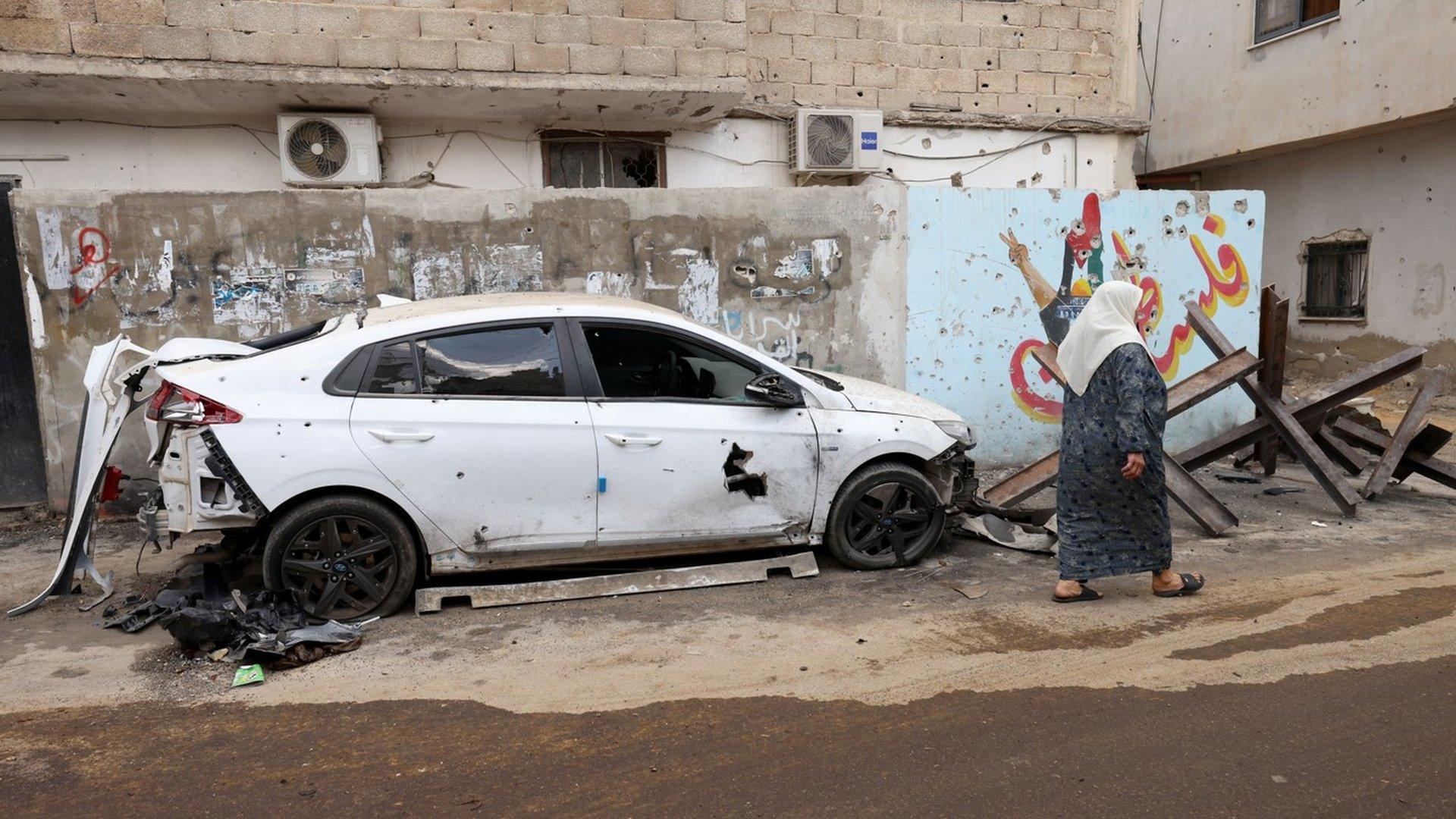 A woman walks past a damaged car in Jenin refugee camp in the occupied West Bank, following a deadly raid by Israeli forces (20 September 2023)