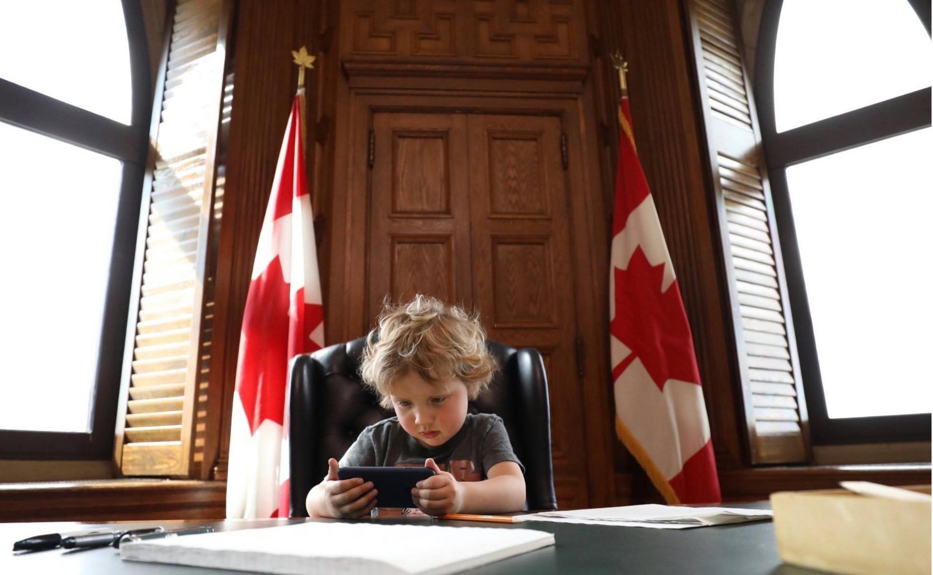 Trudeau junior sits at the prime minister's desk playing with a smartphone, flanked by two Canadian flags