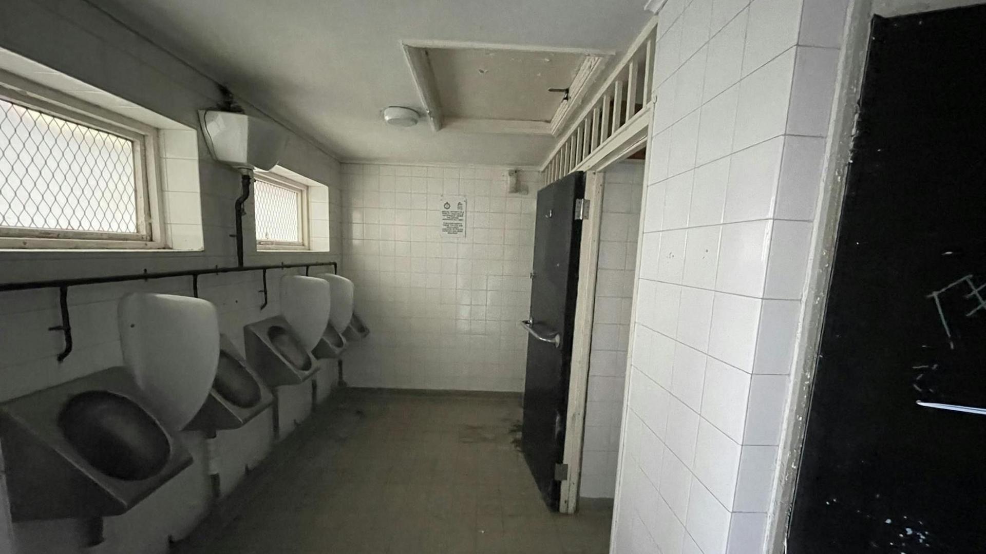 Five metal urinals on a wall on the left of the image. There are two rectangle windows above them. The walls are white tiles and the floor is light brown tiles. there are two black doors on the right wall