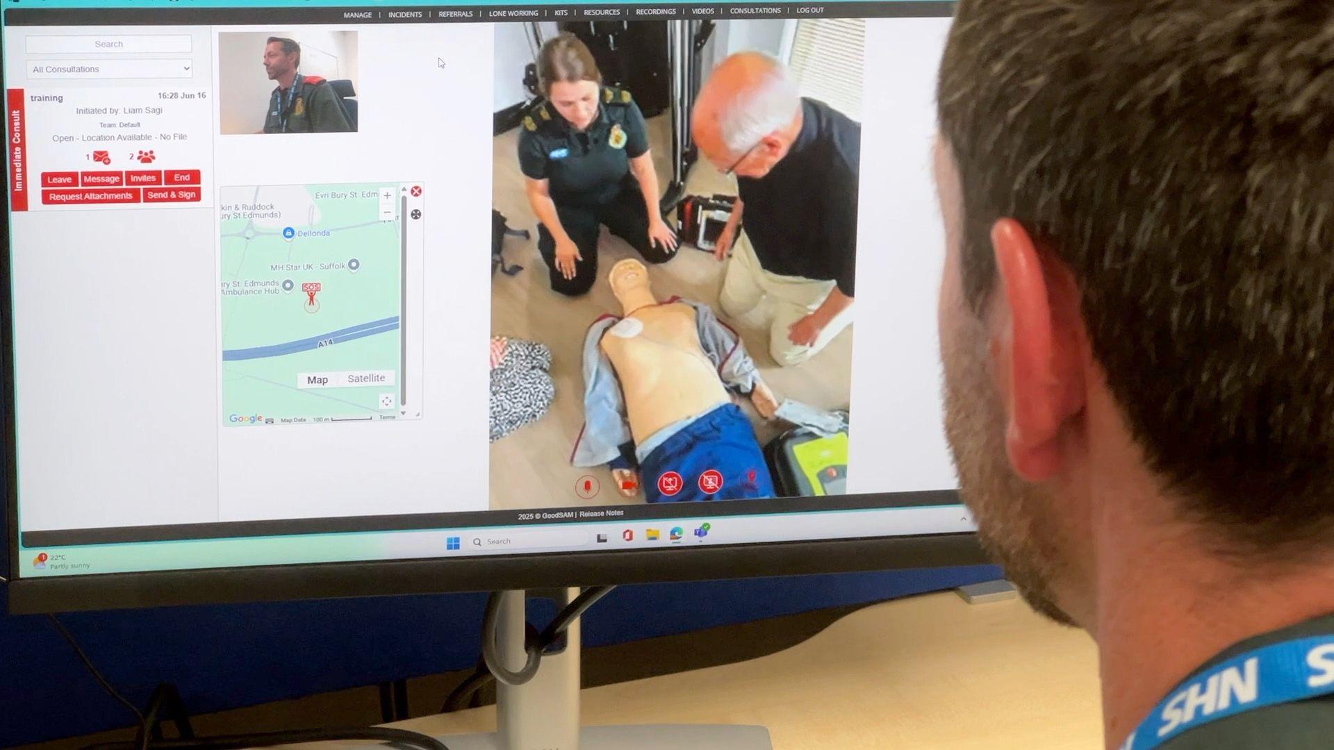 An advanced paramedic looks at his computer screen and sees the moment when ambulance staff arrive to help a bystander who has witnessed someone have a cardiac arrest and has been doing CPR. He is looking at the screen with his head and shoulders and NHS lanyard showing to the right of screen. The computer screen with the image of the dummy patient and bystander is on the left. The computer also shows a map of the location where the patient is and an image of himself.