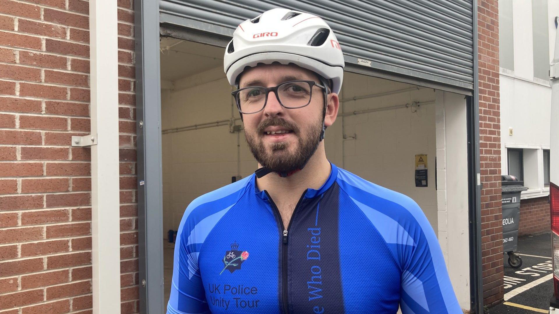 A man is looking at the camera, wearing a cycling helmet and glasses. He has brown hair, moustache, and bears, and is wearing a long sleeved blue cycling shirt which says UK Police Unity Tour. There is an open garage visible behind him.