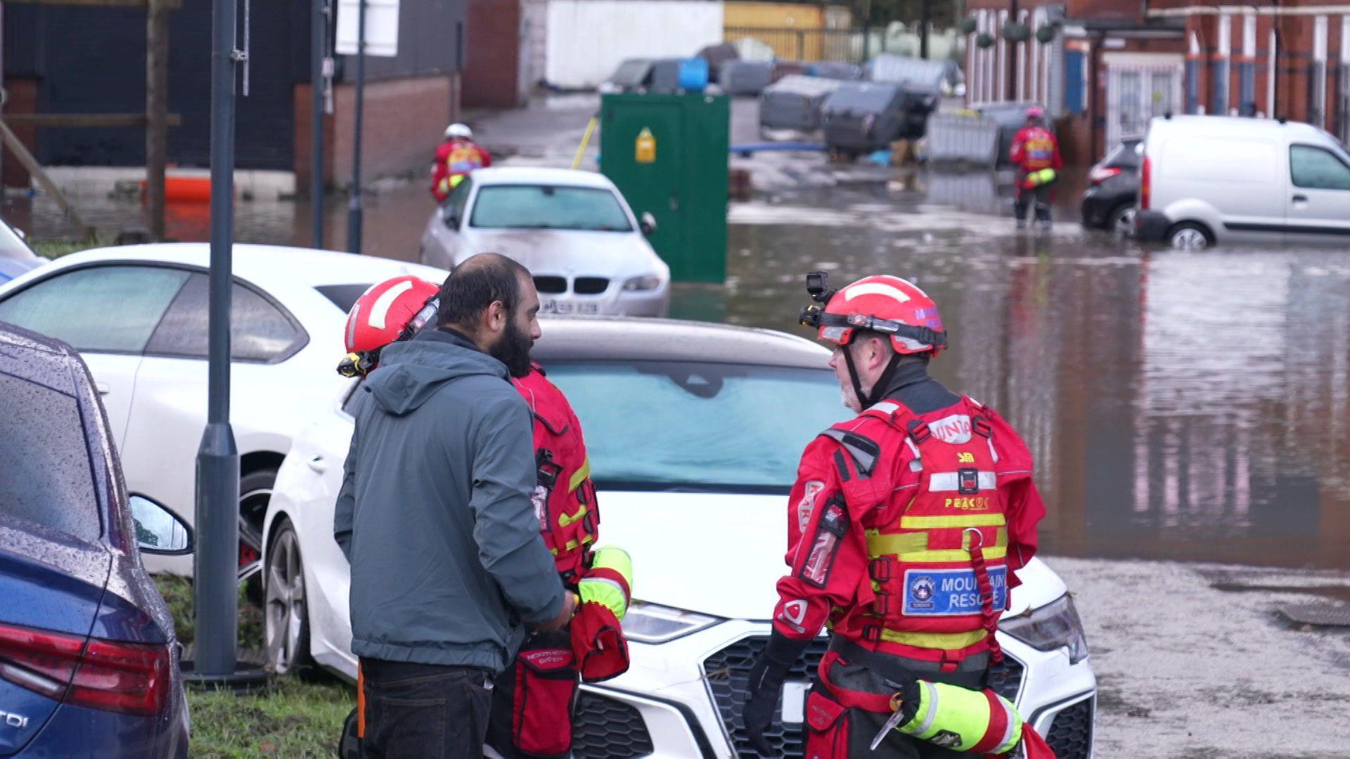 A mountain rescue volunteer in red uniform and wearing a helmet speaks to a man in a flooded street