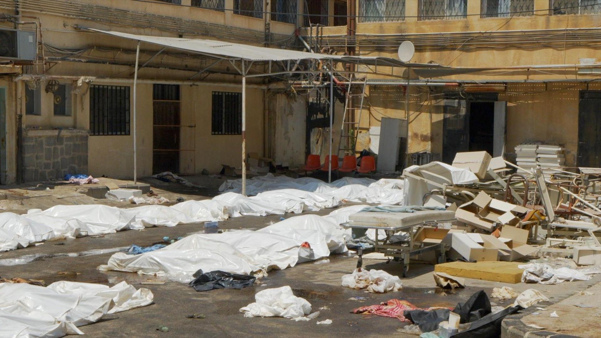 White plastic sheets cover bodies in the carpark of a hospital in Suweida. There is debris of chairs and hospital beds scattered across the ground too