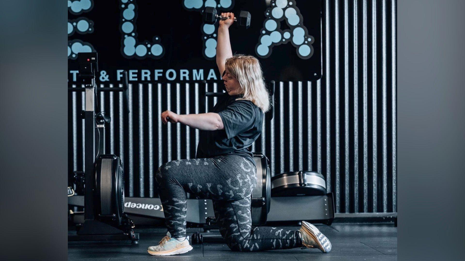 Aisling lifting weights. She is in a gym in leggings and a t-shirt. She has one knee on the floor and the other bent with her foot on the ground. She holds a weight above her head in her right hand.