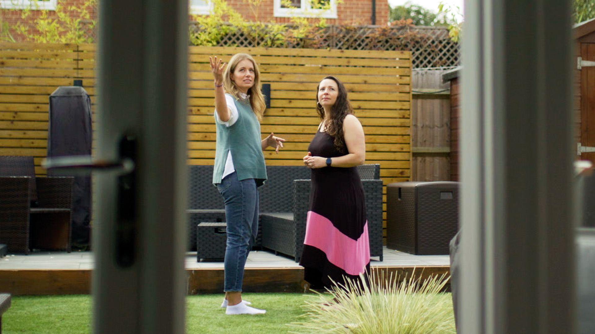 Two women standing and talking in a garden area, viewed through a glass door. The garden has a wooden deck with dark outdoor furniture, a wooden fence with lattice on top, and green grass in the foreground. There is a plant with spiky leaves near the bottom of the image, and a brick building partially visible in the background.