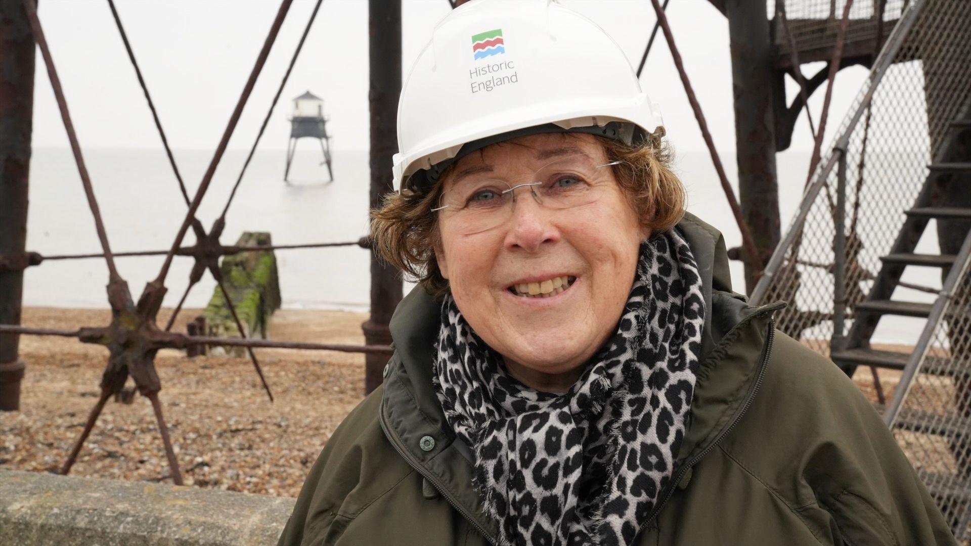 Trudi Hughes from Historic England, standing next to one of the Dovercourt lighthouses, with the second lighthouse pictured over her shoulder out at sea. Trudi is wearing a white hard hat with Historic England branding on the front. She is wearing glasses and smiling at the camera. Trudi is wearing a dark green coat with a leopard-print scarf