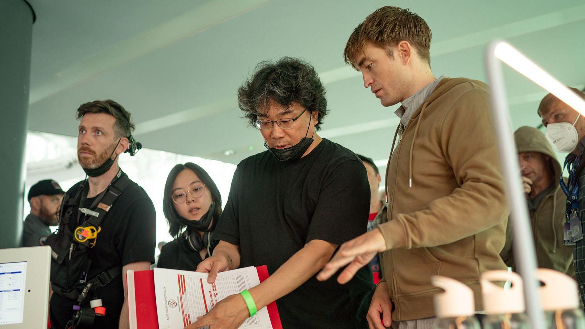Bong Joon Ho and Robert Pattinson speak to one another as one of them holds a sheet of paper while they film Mickey 17.